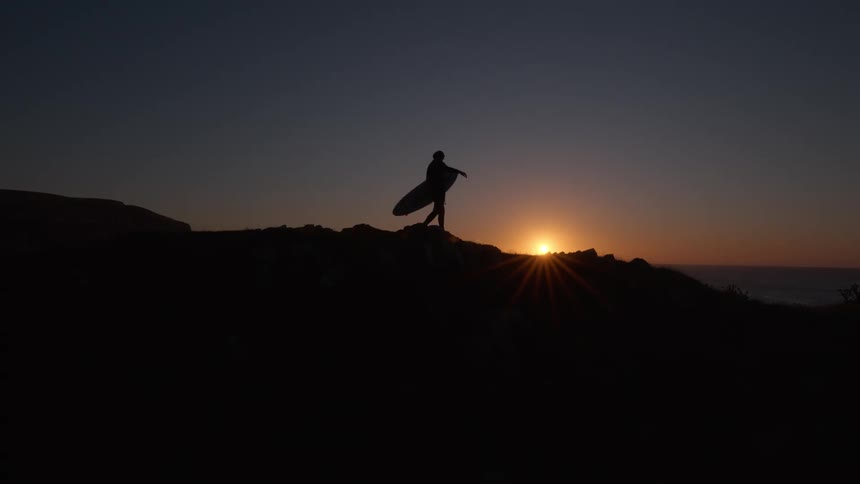 In slow motion, a surfer's silhouette walks along a coastal cliff with a surfboard as the sun sets over the ocean