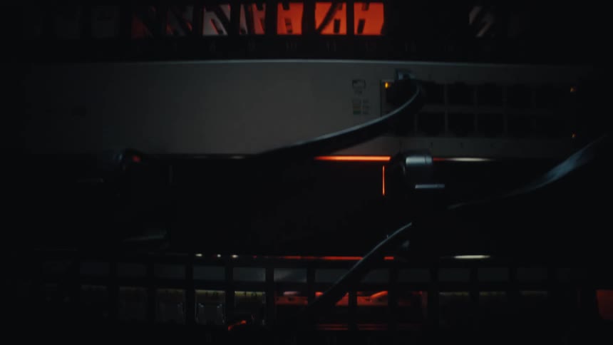 A moody close-up shot tilts up on a network server rack with blinking lights and cables in a dark data room