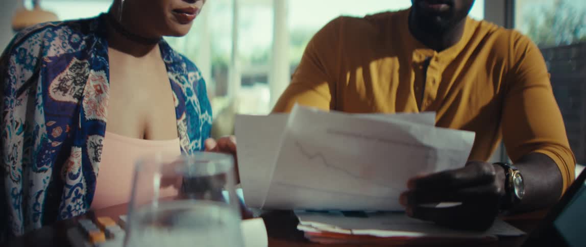 A concerned couple sits at a table discussing their finances while reviewing documents with a downward trending graph