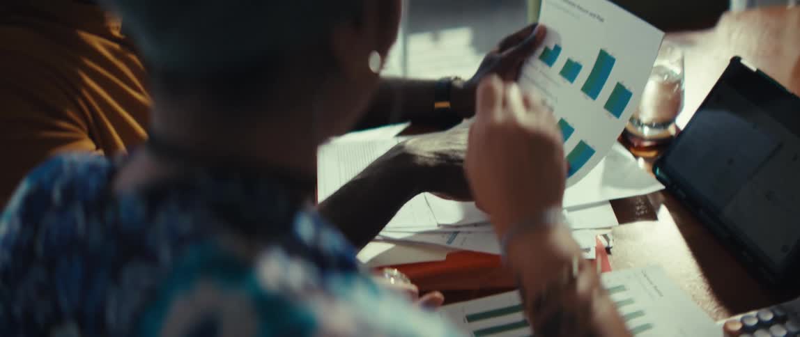 An over-the-shoulder, slow-motion shot of two business colleagues discussing financial data and charts on a printed document during a meeting