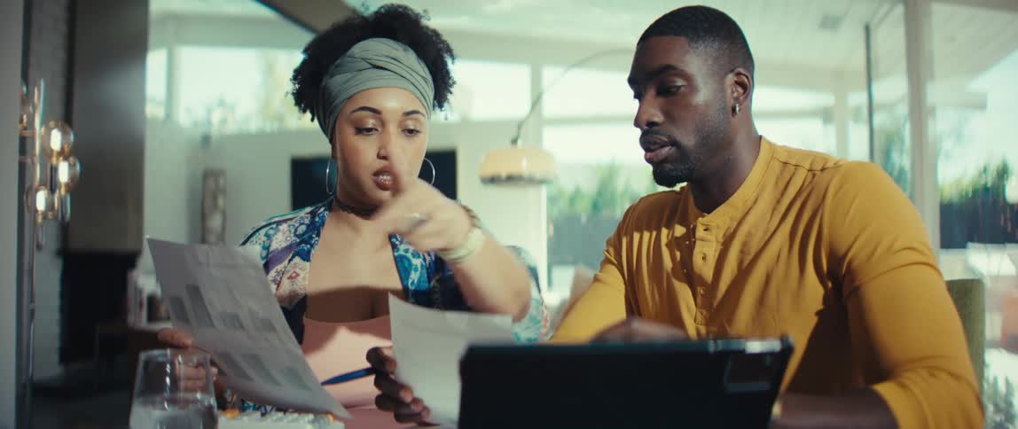 A young Black couple works together on their home finances, using a tablet and reviewing documents at their dining table