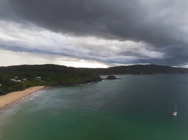 A dramatic aerial pull-out shot reveals a lone sailboat anchored in a scenic bay as massive storm clouds loom overhead