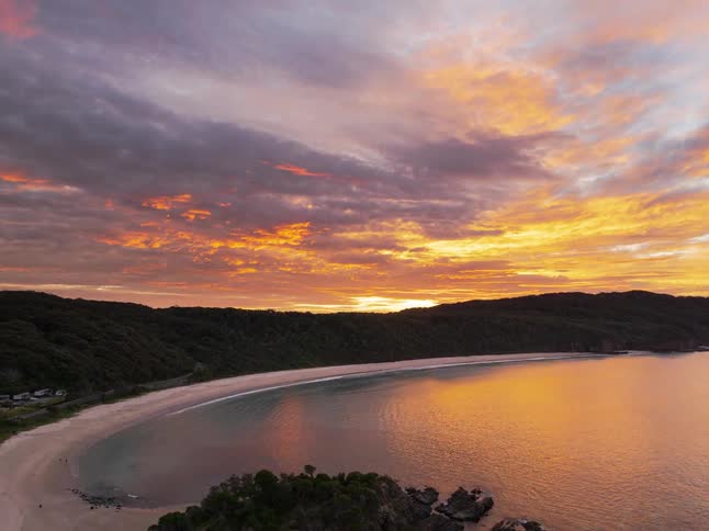 A breathtaking aerial time-lapse captures a fiery red and orange sunset exploding over a tranquil beach and coastal bay