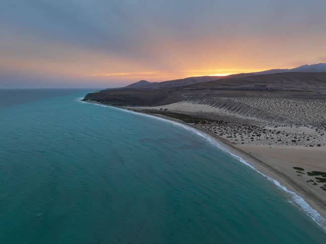 A breathtaking aerial view captures a vast and remote desert coastline meeting the turquoise ocean during a vibrant sunset