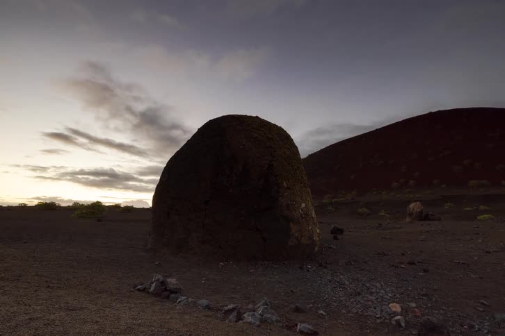 A slow pan reveals a large volcanic boulder resting in a stark, barren landscape near a red cinder cone at dusk