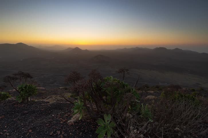 A stunning timelapse captures the sun setting behind a vast volcanic mountain range, with unique desert plants in the foreground