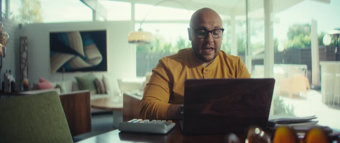 A professional man with glasses works from his modern home office, actively participating in a video call on his laptop