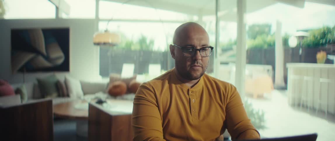 A focused bald man in a yellow shirt and glasses works on his laptop in a modern, bright home