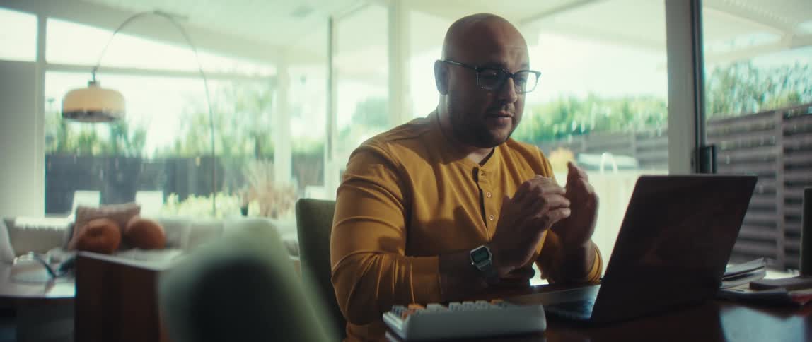 A bald businessman in glasses and a yellow shirt talks and gestures while on a video call in his modern home office