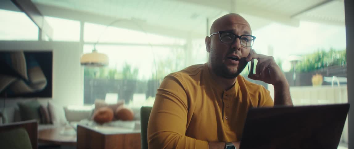 A bald man with glasses in a yellow shirt talks on the phone while working on his laptop in his stylish, sunlit home office