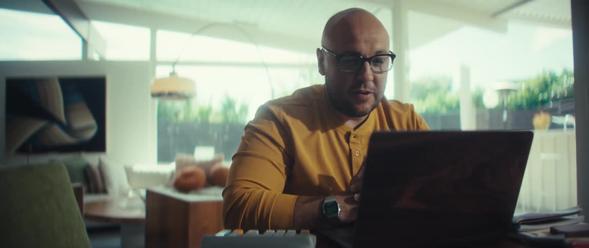 A professional man in a yellow shirt engages in a lively video conference on his laptop from his modern home office