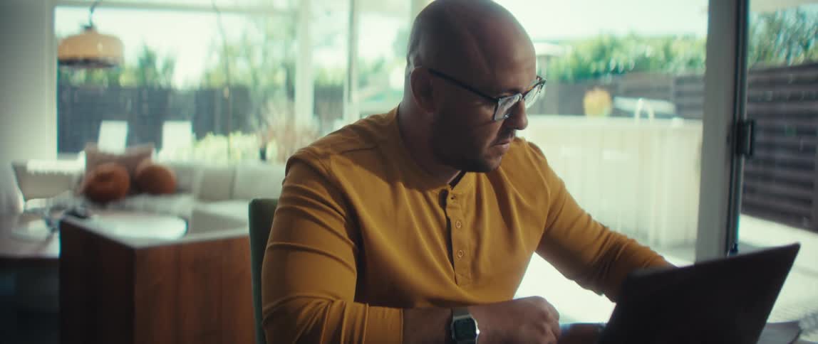 A bald man wearing glasses focuses intently on his laptop while working from his dining room table in a bright, modern home