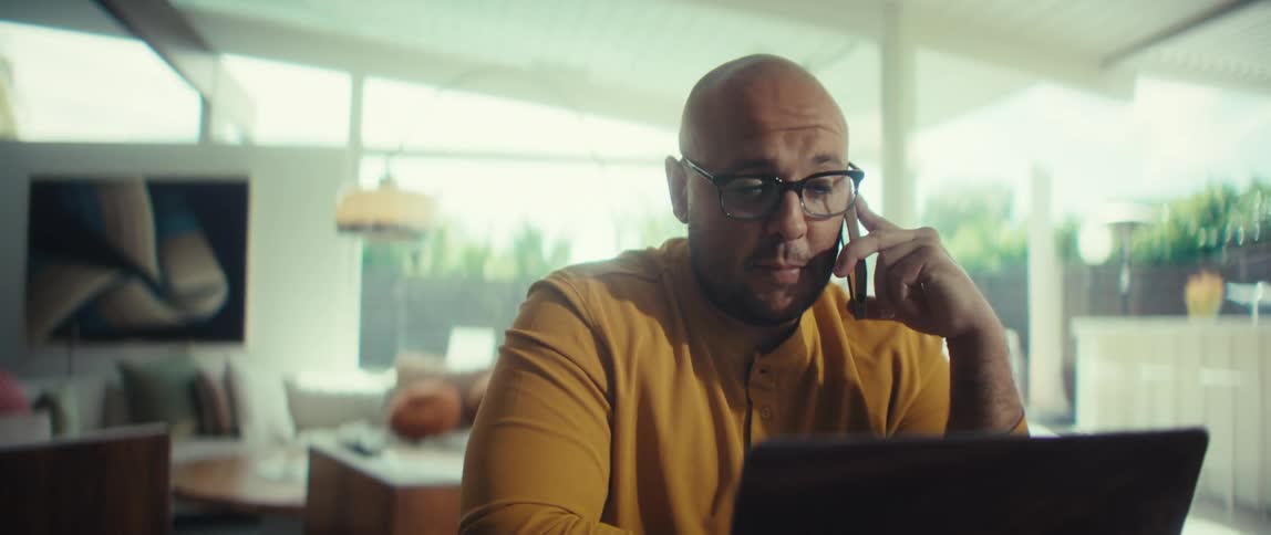 A bald man in a yellow shirt has a serious business call on his phone while working on his laptop in a modern, sunlit home