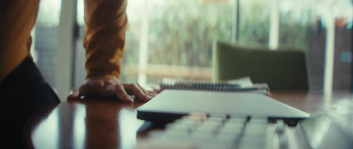 A person's hands finish typing on a laptop, close the lid, and leave the desk in a bright room