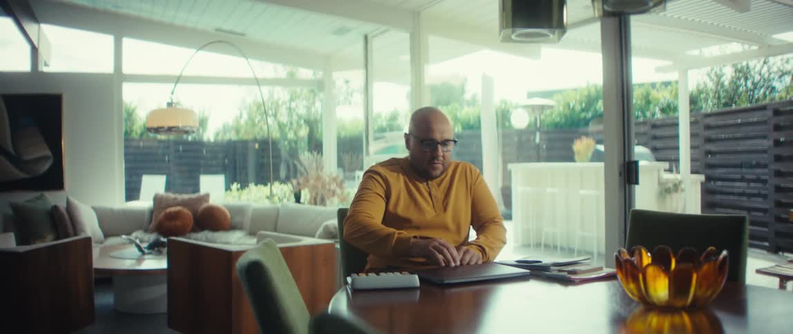 A man in a yellow shirt walks to a table in his stylish modern home, opens his laptop, and begins to work