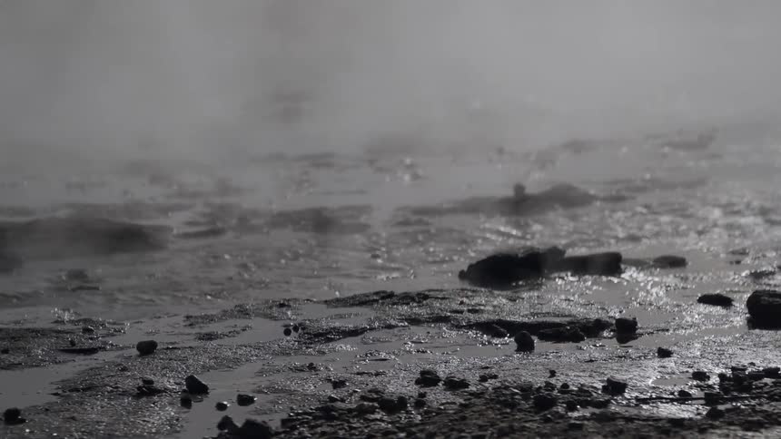 Steam rises from the bubbling water of a geothermal hot spring in a desolate, rocky landscape