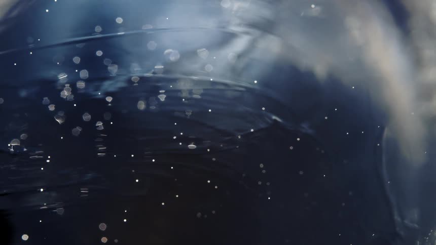 An ethereal macro shot of shimmering particles floating and white smoke swirling in a dark, rippling liquid in slow motion