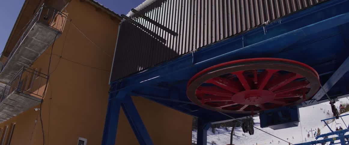 A low-angle shot shows the large red wheel of a chairlift mechanism turning at a sunny ski resort
