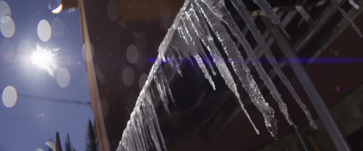 A low angle, slow-motion shot shows icicles melting and dripping from a roof in the bright winter sun