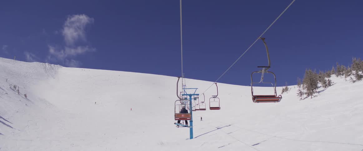 A colorful ski lift carries skiers and snowboarders up a vast, snow-covered mountain under a brilliant blue sky