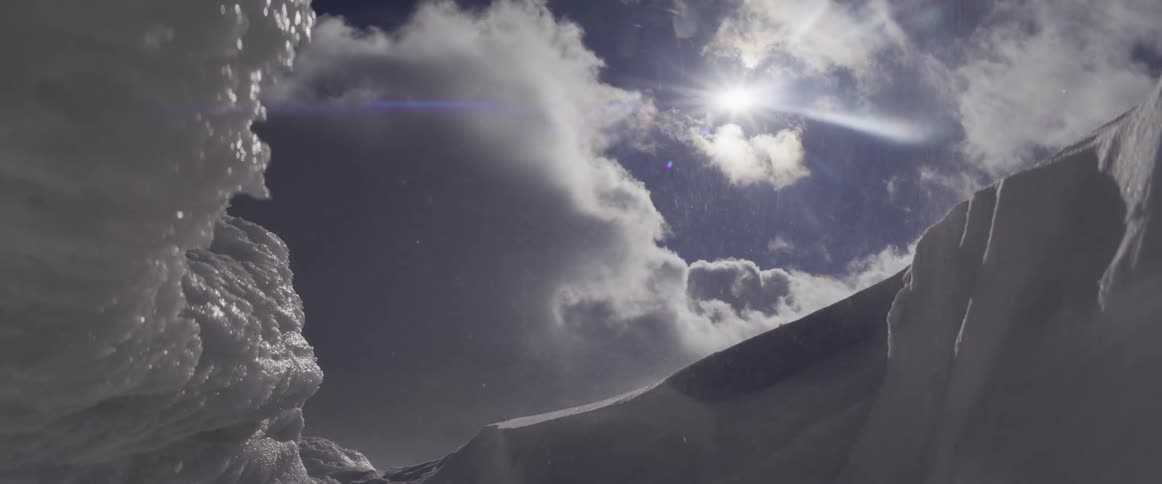 Snow blows across a frozen mountain landscape as the bright sun shines through dramatic clouds