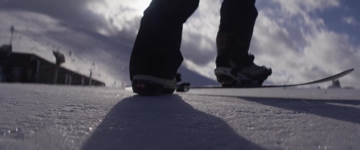 A snowboarder straps into their bindings in slow motion on a glistening, snowy mountain top under a bright sun
