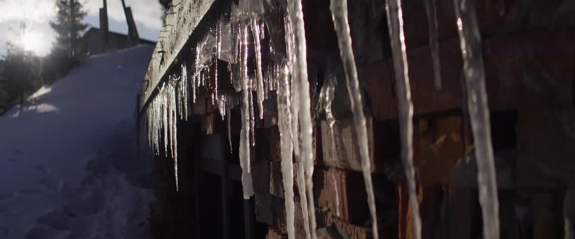 Sunlight glistens on long, sharp icicles hanging from a brick building's roof on a snowy winter day