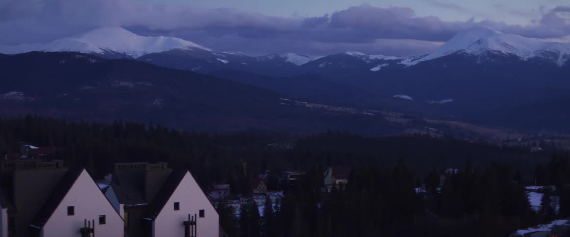 A serene view of snow-capped mountains towering over a forested valley village at dusk