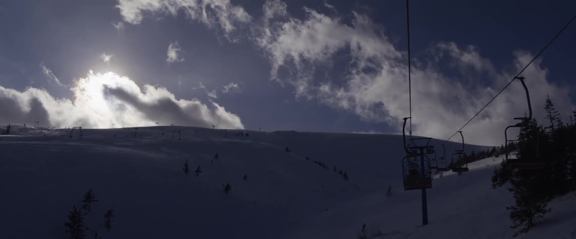 A silhouetted chairlift ascends a snowy mountain under a dramatic, cloudy sky with the sun peeking through