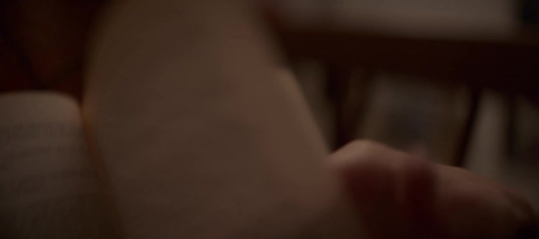 A woman's hand with black nail polish and a silver ring delicately turns the page of a book in a cozy setting
