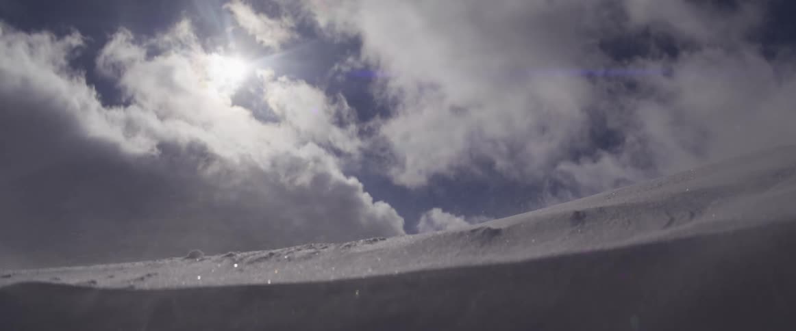 Glistening snow blows across a vast snowy landscape in slow motion as the sun peaks through the clouds