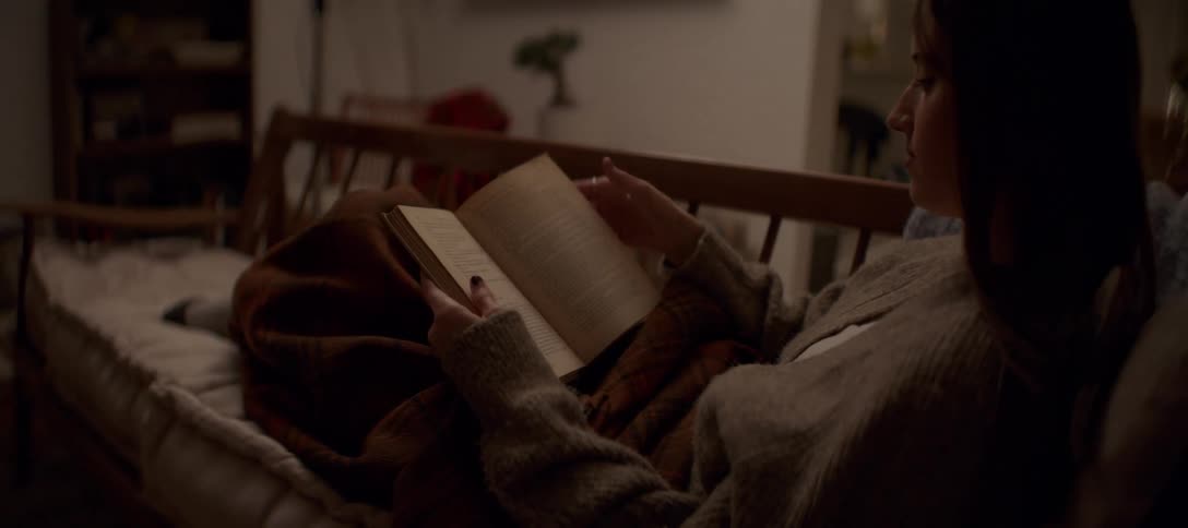 A cozy young woman flips through the pages of an old book while relaxing on a couch at night