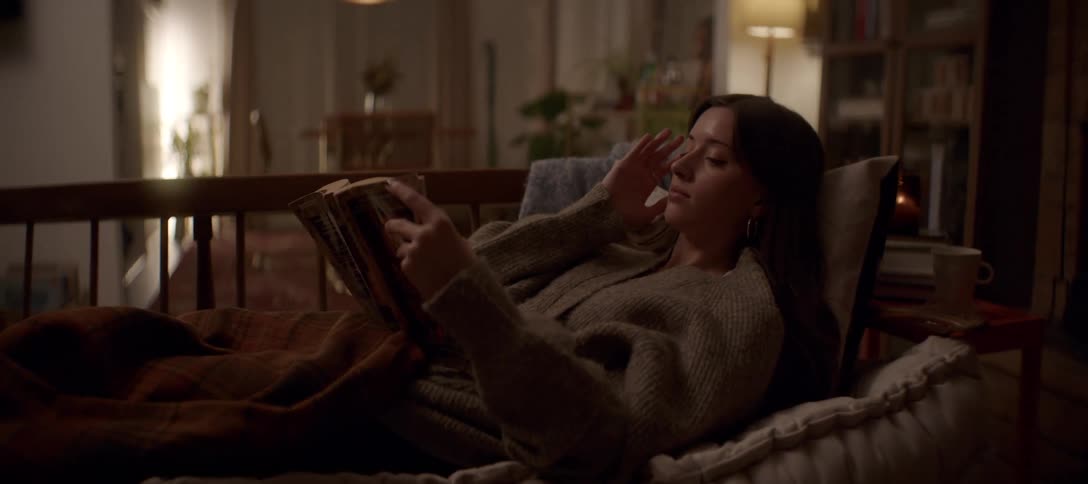 A woman relaxes on a cozy couch, engrossed in a book in her warmly lit living room in the evening