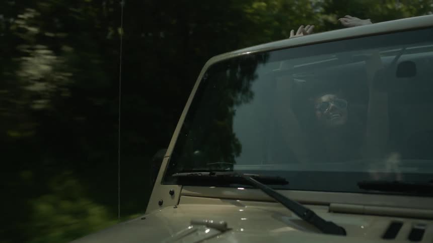 A woman rides in the passenger seat of a jeep with her hands raised through the sunroof