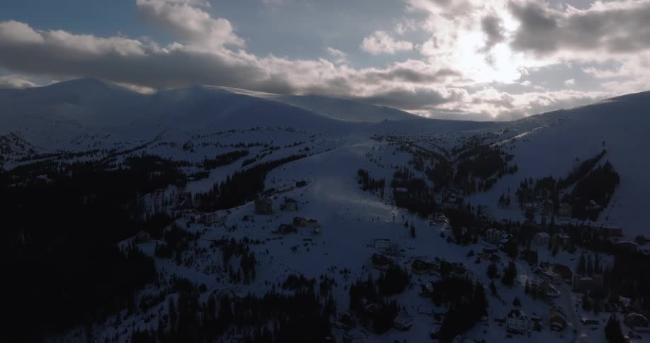 A majestic aerial view reveals a quiet ski resort village nestled in a vast, snow-covered mountain valley under a dramatic, cloudy sky