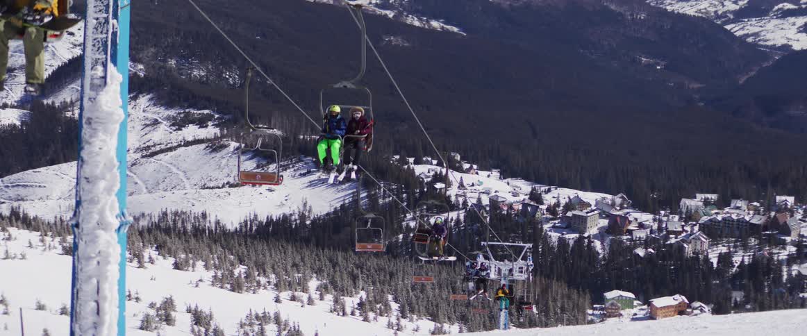 Skiers and snowboarders ride a chairlift up a snow-covered mountain overlooking a bustling ski resort village
