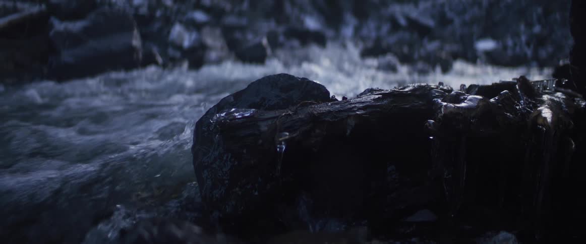 Cool water rushes past a dark, wet rock in a riverbed in a cinematic slow-motion shot