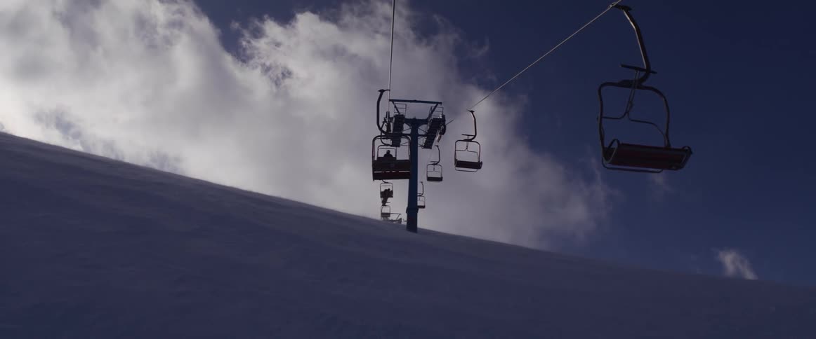 A silhouetted ski lift carries people up a vast, snowy mountain slope as wind blows snow against a cloudy sky