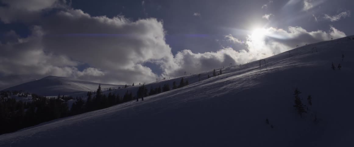Sunlight shines over a shadowy, snow-covered mountainside at a ski resort under a cloudy sky
