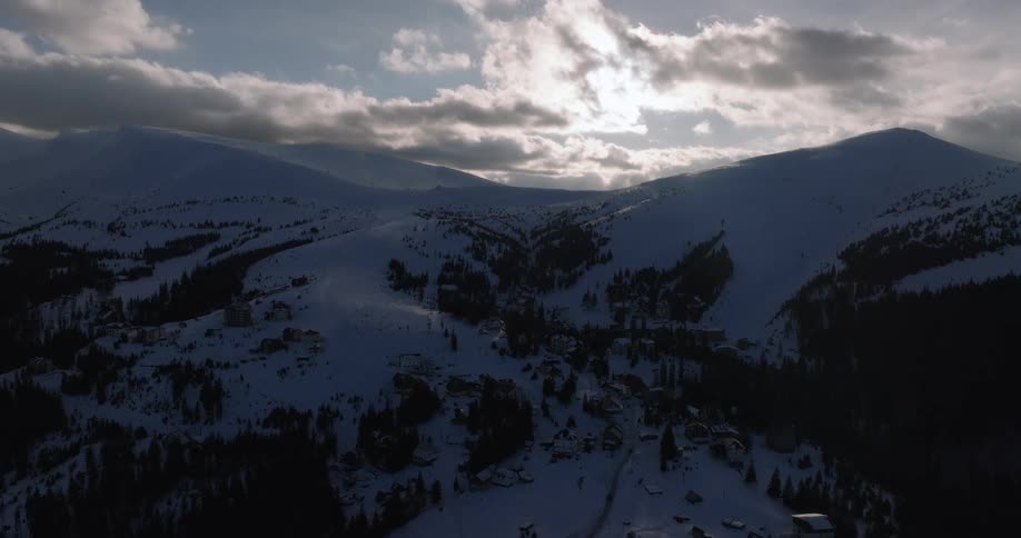Slow aerial reveal of a remote village tucked away in a snowy mountain valley under a dramatic, cloudy sky