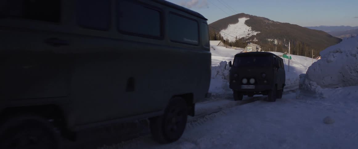 Two rugged off-road vans drive in a convoy through a remote, snow-covered mountain village on a clear winter day