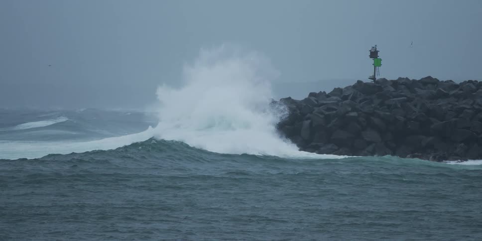 A powerful ocean wave crashes against a rock jetty in slow motion during a storm
