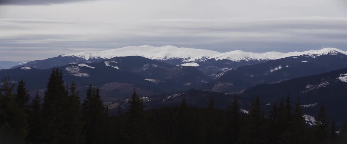 A slow pan across a majestic winter landscape reveals a snow-capped mountain range rising above a dense evergreen forest