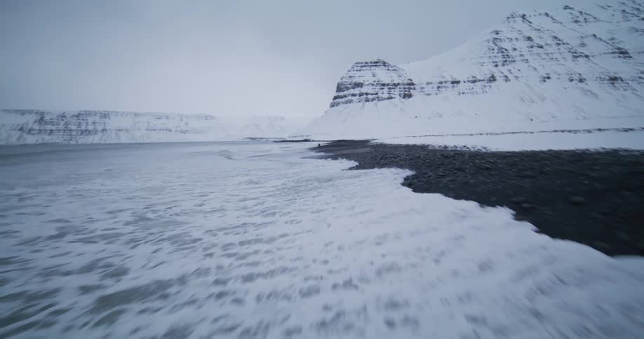 A slow-motion aerial shot follows the frigid ocean waves crashing on a snowy beach, revealing a lone surfer walking along the shore