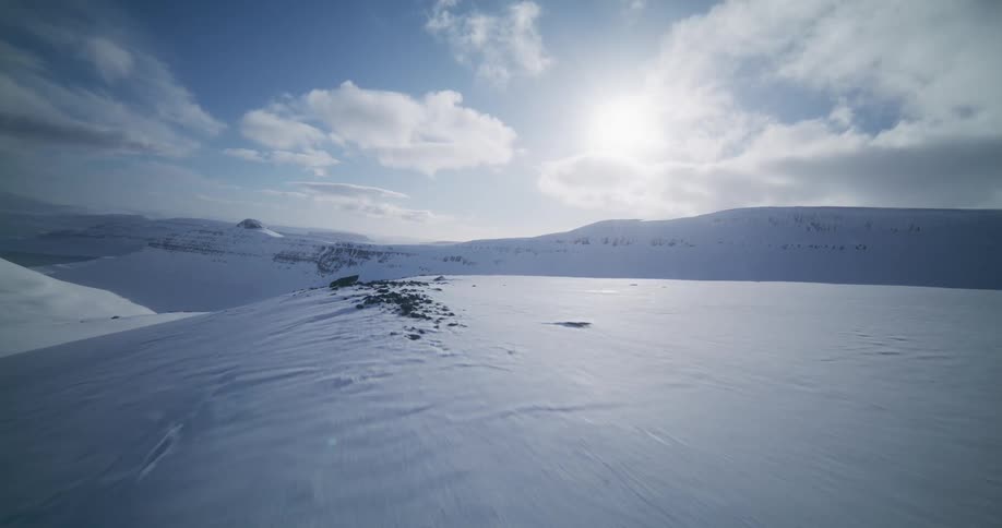 A breathtaking first-person aerial view soars low and fast over a vast, sun-drenched, snow-covered mountain landscape