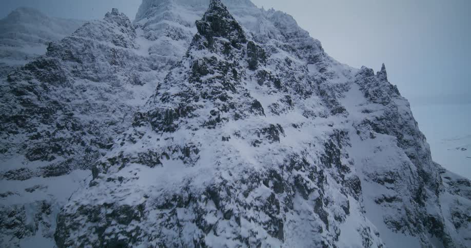 A dramatic aerial view slowly reveals the immense scale of a rugged, snow-covered mountain peak