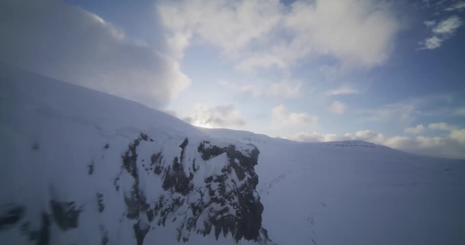 A dramatic FPV drone flight soars along the edge of a vast, snow-covered mountain landscape under a bright, cloudy sky