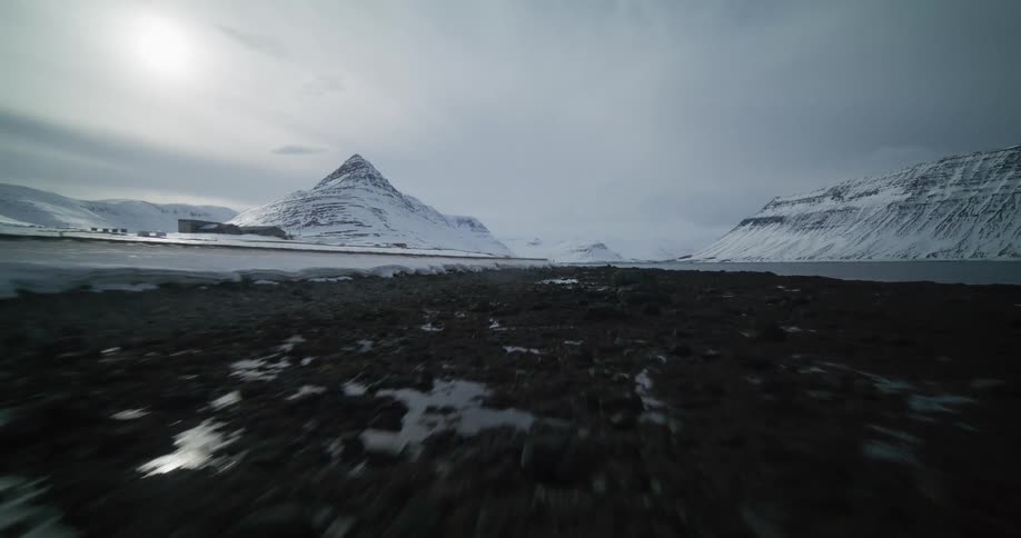 A low, fast aerial shot glides over the rocky, icy shoreline towards the snow-covered Kirkjufell mountain in Iceland