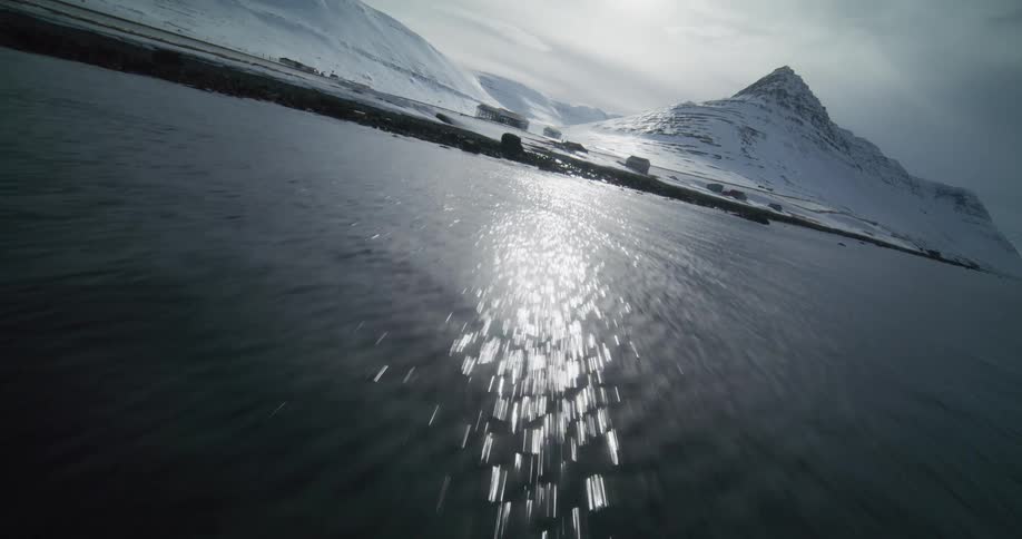 A fast-paced aerial shot skims low over the sun's reflection on a fjord's surface towards a snowy, mountainous shoreline in winter