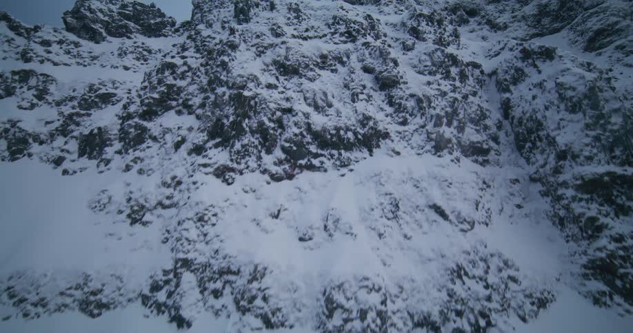 An aerial ascent up a steep and rugged snow-covered mountain face reveals a vast and desolate winter landscape